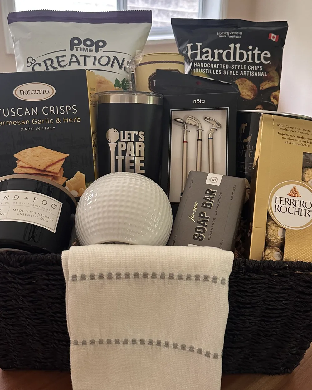 Golf-themed gift basket with golf balls, snacks, chocolate and a striped towel in a dark woven basket